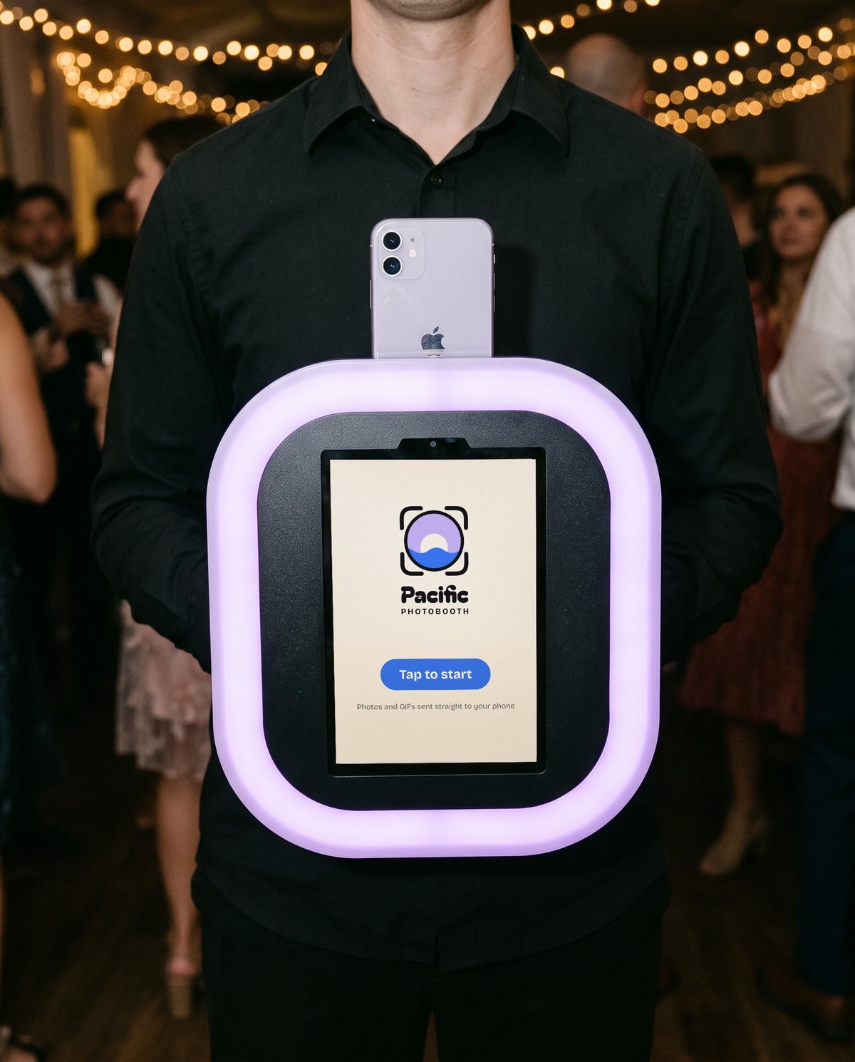 A Pacific Photobooth photographer in a black shirt holding the roaming booth at a party: an iPad showing Tap to start in a glowing lilac frame